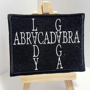 May include: Black fabric patch with white embroidery that reads "L G ABRVCADAVBRA D Y G A". The patch is displayed on a small wooden easel.