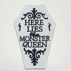 Puede incluir: Parche blanco en forma de ata&uacute;d con texto bordado en negro y adornos decorativos. El texto dice "HERE LIES the MONSTER QUEEN". El parche tiene un borde blanco, para ropa o accesorios.