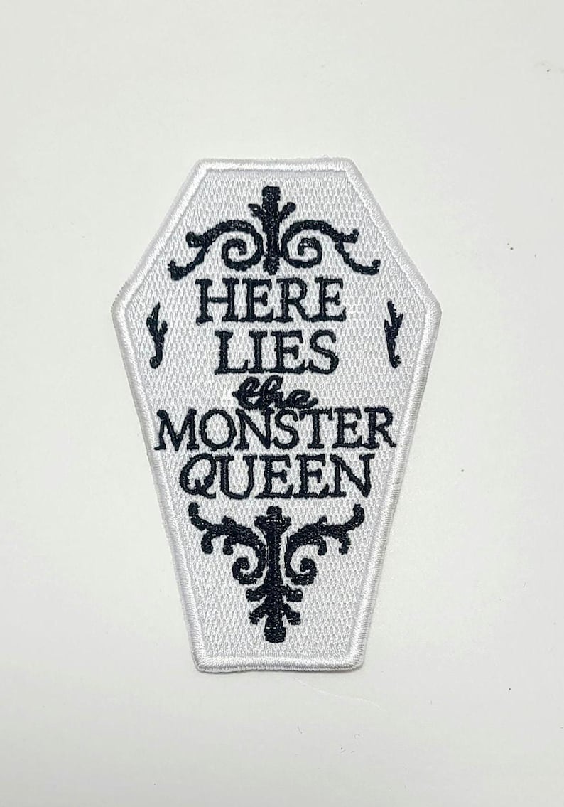 Puede incluir: Parche blanco en forma de ata&uacute;d con texto bordado en negro y adornos decorativos. El texto dice "HERE LIES the MONSTER QUEEN". El parche tiene un borde blanco, para ropa o accesorios.