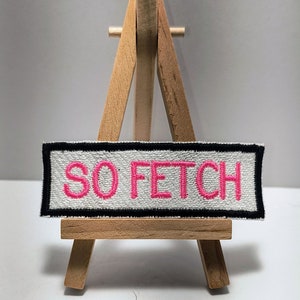 May include: A white fabric patch with black border and pink embroidered text "SO FETCH" displayed on a small wooden easel.