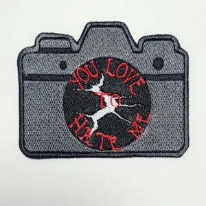 Può includere: Toppa ricamata a forma di macchina fotografica grigia. Un cerchio nero centrale riporta la scritta rossa "YOU LOVE TO HATE". La toppa è posta su sfondo bianco.