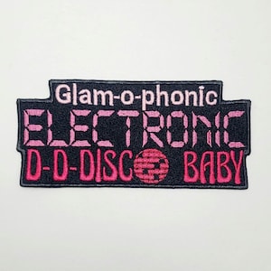 May include: A black patch with pink embroidered text that reads "Glam-o-phonic ELECTRONIC D-D-DISCO BABY". The patch has a rectangular shape with a unique top edge and a pink and black color scheme.