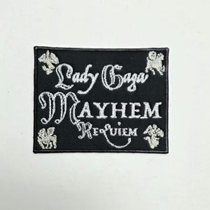 May include: Black rectangular patch with white embroidered text reading "Lady Gaga Mayhem Requiem." The patch has decorative embroidered accents in each corner. The patch is likely for a jacket or bag.