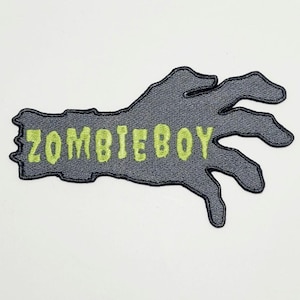 May include: A gray embroidered patch in the shape of a reaching hand. The word "ZOMBIEBOY" is embroidered in bright green across the hand. The patch has a black border and is set against a white background.