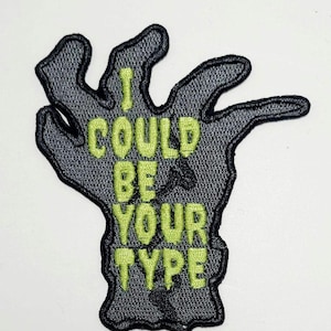 Op de afbeelding: Geborduurde patch in de vorm van een hand met de tekst "I COULD BE YOUR TYPE" in neon groen. De hand is grijs met een zwarte omtrek, en de tekst is in een vetgedrukte, schreefloze lettertype. De patch is waarschijnlijk voor kleding of accessoires.