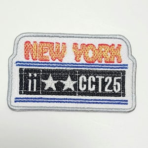 Peut inclure: Écusson brodé avec une bordure blanche et une forme rectangulaire. Le haut de l'écusson indique "NEW YORK" en orange et jaune. En dessous, un rectangle noir présente deux étoiles blanches et le texte "CCT25".