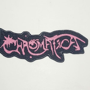 Pode incluir: Patch bordado com a palavra "CHROMATICA" em rosa, com uma tipografia estilizada. O patch tem um fundo preto e uma forma irregular. Ideal para roupas ou acessórios.