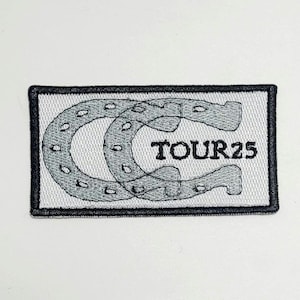 May include: Embroidered patch with a black border and a white background. The patch features a gray horseshoe design with the text "TOUR25" in black. The patch is rectangular in shape.