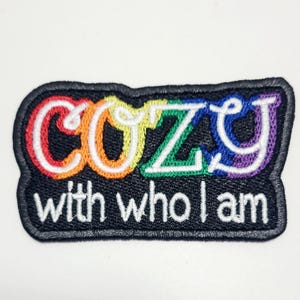 Könnte beinhalten: Ein schwarzer, gestickter Aufnäher mit dem Wort "COZY" in Regenbogenfarben und dem Schriftzug "with who I am" in Weiß. Der Aufnäher hat einen schwarzen Rand und ist für Kleidung oder Accessoires gedacht.