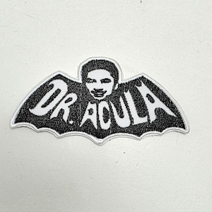 May include: Black and white embroidered patch featuring the text "DR. ACULA" in a stylized font. The design includes a bat-wing shape with a portrait of a person in the center. The patch has a white border.