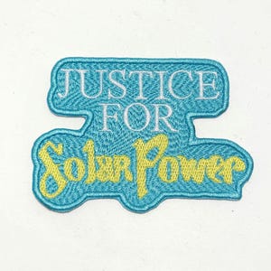 May include: Embroidered patch in a teal blue color with the words "JUSTICE FOR" in white and "Solar Power" in yellow. The patch has a unique shape with a teal border.