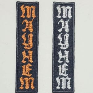 Può includere: Due toppe rettangolari con la parola "MAYHEM" ricamata verticalmente in un carattere gotico. Una toppa presenta filo arancione su sfondo nero, l'altra filo bianco. Entrambe hanno un bordo nero.