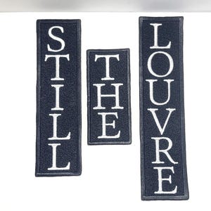 May include: Three rectangular patches with white embroidered text on a dark blue background. The patches read "STILL", "THE", and "LOUVRE" in a vertical arrangement. The patches are of varying sizes.