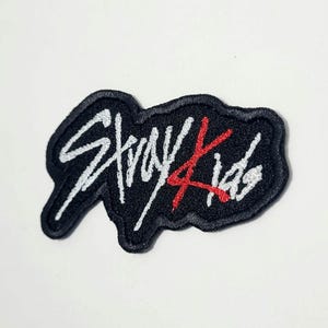 Puede incluir: Parche bordado con el texto "Stray Kids" en escritura blanca y detalles rojos sobre un fondo negro. El parche tiene una forma irregular única y un borde negro.