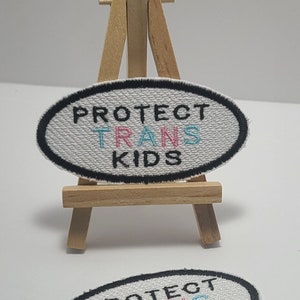 May include: White oval patch with black border and the text "PROTECT TRANS KIDS" in black, pink, and blue lettering. The patch is displayed on a small wooden easel.