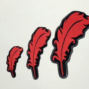 Peut inclure: Trois écussons de plumes rouges avec des bordures noires et grises sont présentés sur un fond blanc. Les écussons sont de tailles différentes, le plus grand étant à droite. Les plumes ont des motifs de veines détaillés.