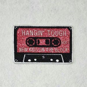 May include: A rectangular embroidered patch shaped like a cassette tape. The top section is red with a glittery texture, featuring the white text HANGIN' TOUGH and NEW KIDS ON THE BLOCK. The bottom section is black with white detailing.