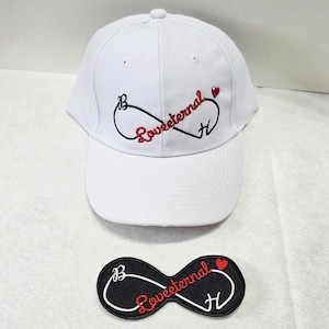 May include: A white baseball cap featuring an embroidered black infinity symbol, the red script text "Loveeternal", a small red heart, and the letters B and H. Below the cap is a matching black patch with the same embroidered design.