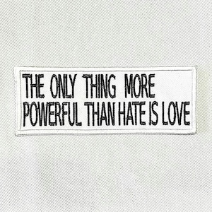 Puede incluir: Un parche rectangular blanco con texto bordado en negro: "THE ONLY THING MORE POWERFUL THAN HATE IS LOVE". El parche tiene un borde negro y está sobre un fondo de tela gris claro.