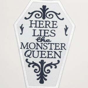 Puede incluir: Parche blanco en forma de ata&uacute;d con texto negro que dice "HERE LIES the MONSTER QUEEN". El parche tiene un borde blanco y adornos negros decorativos. Una cinta m&eacute;trica est&aacute; en la parte inferior.