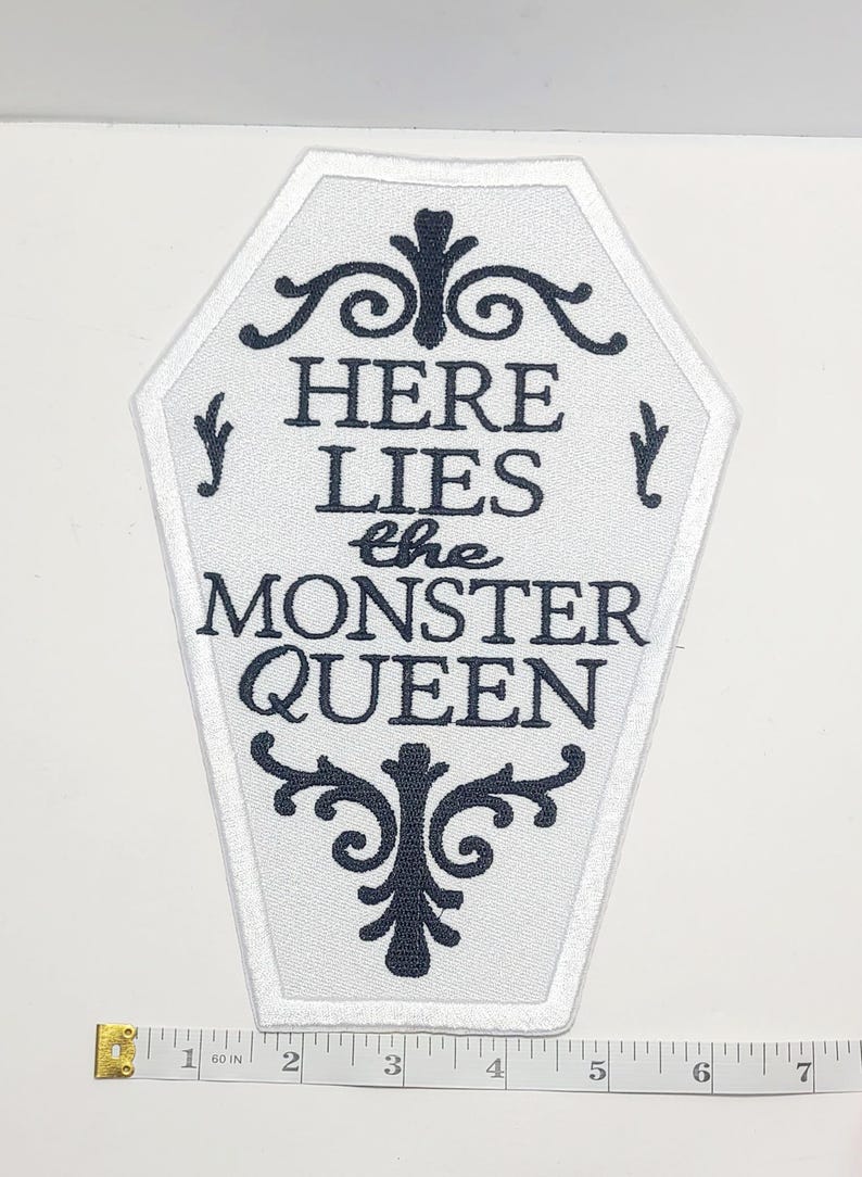 Puede incluir: Parche blanco en forma de ata&uacute;d con texto negro que dice "HERE LIES the MONSTER QUEEN". El parche tiene un borde blanco y adornos negros decorativos. Una cinta m&eacute;trica est&aacute; en la parte inferior.