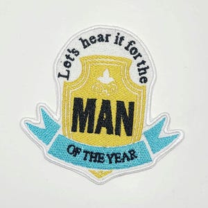 May include: Embroidered patch with a yellow and black shield design. The patch features the words "Let's hear it for the MAN of the YEAR" in black and white lettering, with a light blue banner at the bottom.