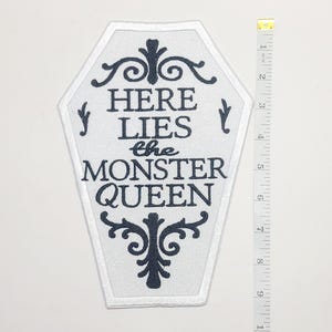 Puede incluir: Un parche blanco en forma de ata&uacute;d con texto negro que dice "HERE LIES the MONSTER QUEEN". El parche tiene un borde blanco y adornos negros decorativos. Una cinta m&eacute;trica amarilla y blanca est&aacute; a la derecha del parche, mostrando pulgadas.