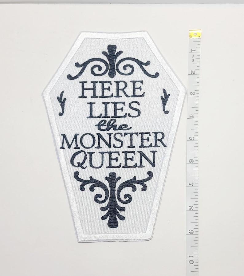 Puede incluir: Un parche blanco en forma de ata&uacute;d con texto negro que dice "HERE LIES the MONSTER QUEEN". El parche tiene un borde blanco y adornos negros decorativos. Una cinta m&eacute;trica amarilla y blanca est&aacute; a la derecha del parche, mostrando pulgadas.