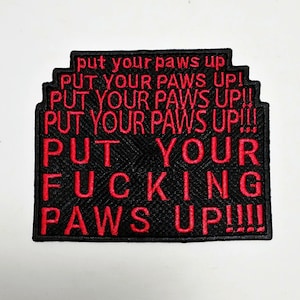 May include: Black embroidered patch with red text that reads "put your paws up" multiple times, including "PUT YOUR FUCKING PAWS UP!!!". The patch has a textured background and a stepped border.