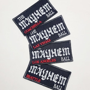 Op de afbeelding: Vier zwarte geborduurde patches met de tekst "THE MAYHEM BALL" in witte gotische letters. Elke patch heeft een andere stadsnaam in het rood: Coachella, Las Vegas, Los Angeles en Seattle.