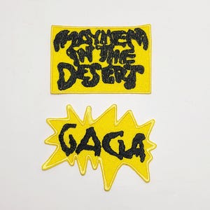 Puede incluir: Dos parches bordados con fondos amarillos y costuras negras. El parche rectangular superior presenta el texto "MAYHEM IN THE DESERT" en una fuente estilizada. El parche inferior tiene forma de estrella dentada y muestra el texto "GAGA".