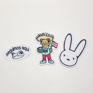 Puede incluir: Tres parches bordados sobre fondo blanco. Uno presenta una rana de dibujos animados con el texto "seguimos aqui". Otro representa un personaje de dibujos animados sosteniendo una cámara y una bandera puertorriqueña. El tercer parche es un conejito blanco con ojos en forma de cruz negros.