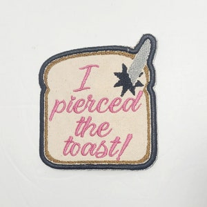 May include: A beige embroidered patch shaped like a slice of bread with a knife sticking out of the top. The text on the patch reads "I pierced the toast!" in pink thread.