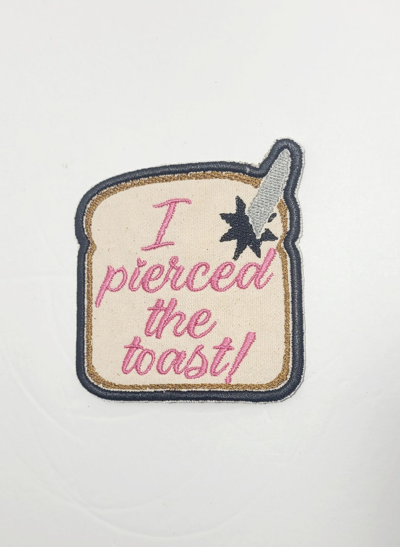 May include: A beige embroidered patch shaped like a slice of bread with a knife sticking out of the top. The text on the patch reads "I pierced the toast!" in pink thread.