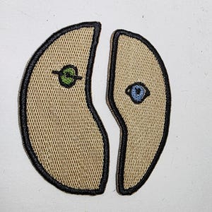 May include: Two embroidered patches, each in a tan color with a black border. One patch features a green eye design, and the other has a blue eye design. The patches are separated, creating a split design.