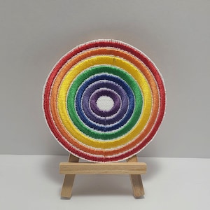 May include: A rainbow-colored embroidered patch with a white background. The patch is circular and has a concentric circle design with red, orange, yellow, green, blue, indigo, and violet colors. The patch is displayed on a small wooden easel.