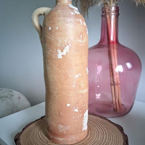 May include: A rustic, terracotta-colored ceramic bottle with a handle. The bottle has a worn, white-painted finish and is sitting on a wooden slice.