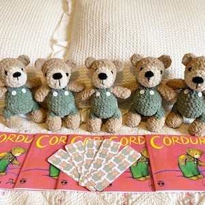 May include: Five handmade crochet teddy bears with tan fur and green overalls are arranged on a bed with matching books and cards. The books have the title "Corduroy" in yellow letters on a pink background.