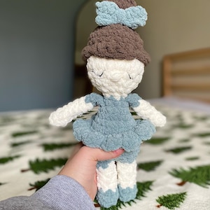 May include: A handmade crochet doll with a white face, closed eyes, and a brown hat with a light blue bow. The doll wears a light blue dress and has white arms and legs with light blue accents. The doll is held in front of a green and white patterned blanket.
