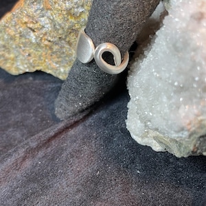 May include: A silver ring with a unique, abstract design. The ring is made of smooth, polished metal and has a modern, minimalist aesthetic.