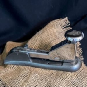 May include: A vintage silver stapler with a black base, resting on a burlap surface.
