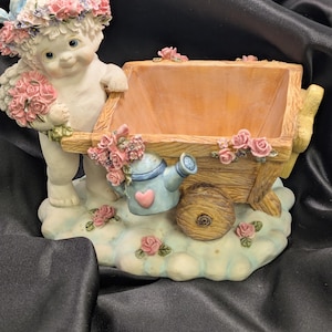 May include: A cherubic figurine pushing a wooden wheelbarrow planter. The cherub wears a floral crown and holds a bouquet of pink roses. A small blue watering can with a heart is attached to the wheelbarrow, which is decorated with more pink roses.