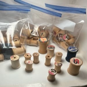 May include: A collection of vintage wooden thread spools in various sizes and colors, including white, brown, blue, and red. Some spools have labels with text.