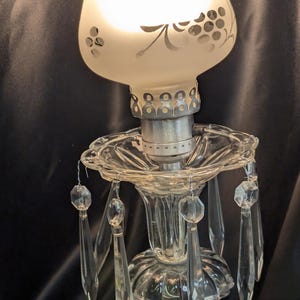 May include: A vintage-style table lamp with a frosted glass shade featuring a floral design. The lamp has a clear glass base with crystal accents and hanging prisms. The lamp is turned on, emitting a soft glow.
