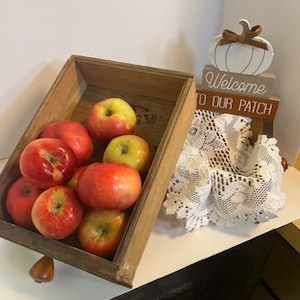 May include: A wooden crate filled with red and yellow apples. A white pumpkin with a brown bow sits on top of a wooden sign that reads "Welcome to our Patch".