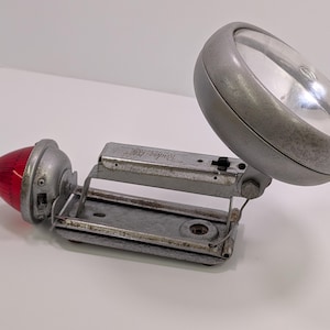 May include: A vintage silver-colored flashlight with a red lens and a folding handle. The flashlight has a clear glass lens and a switch on the side. The text "Radiac" is printed on the side of the flashlight.