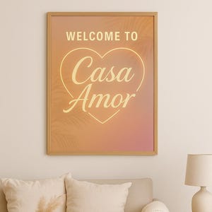 May include: A framed print with the text "WELCOME TO Casa Amor" in a heart shape. The artwork features a warm colour gradient and is displayed above a beige sofa with cushions and a wooden coffee table.