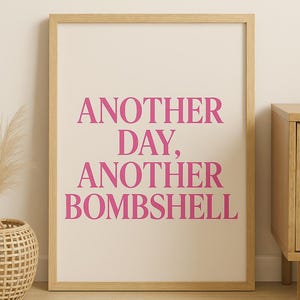 May include: A framed print with the text "ANOTHER DAY, ANOTHER BOMBSHELL" in bright pink. The text is centered on a cream-colored background, within a light wood frame. The print is displayed in a room with light wood furniture and flooring.