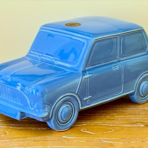 May include: A blue ceramic car figurine, a miniature replica of a classic vehicle. The car has a glossy finish, with details like wheels, windows, and a front grille. The figurine is a solid blue colour and sits on a wooden surface.