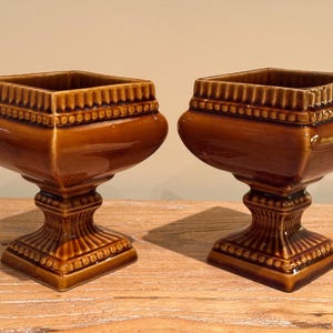 Peut inclure: Deux jardinières en céramique marron assorties, avec un dessus carré et une base en piédestal. Les jardinières ont une finition brillante et des détails décoratifs autour du bord et de la base. Elles sont exposées sur une surface en bois.